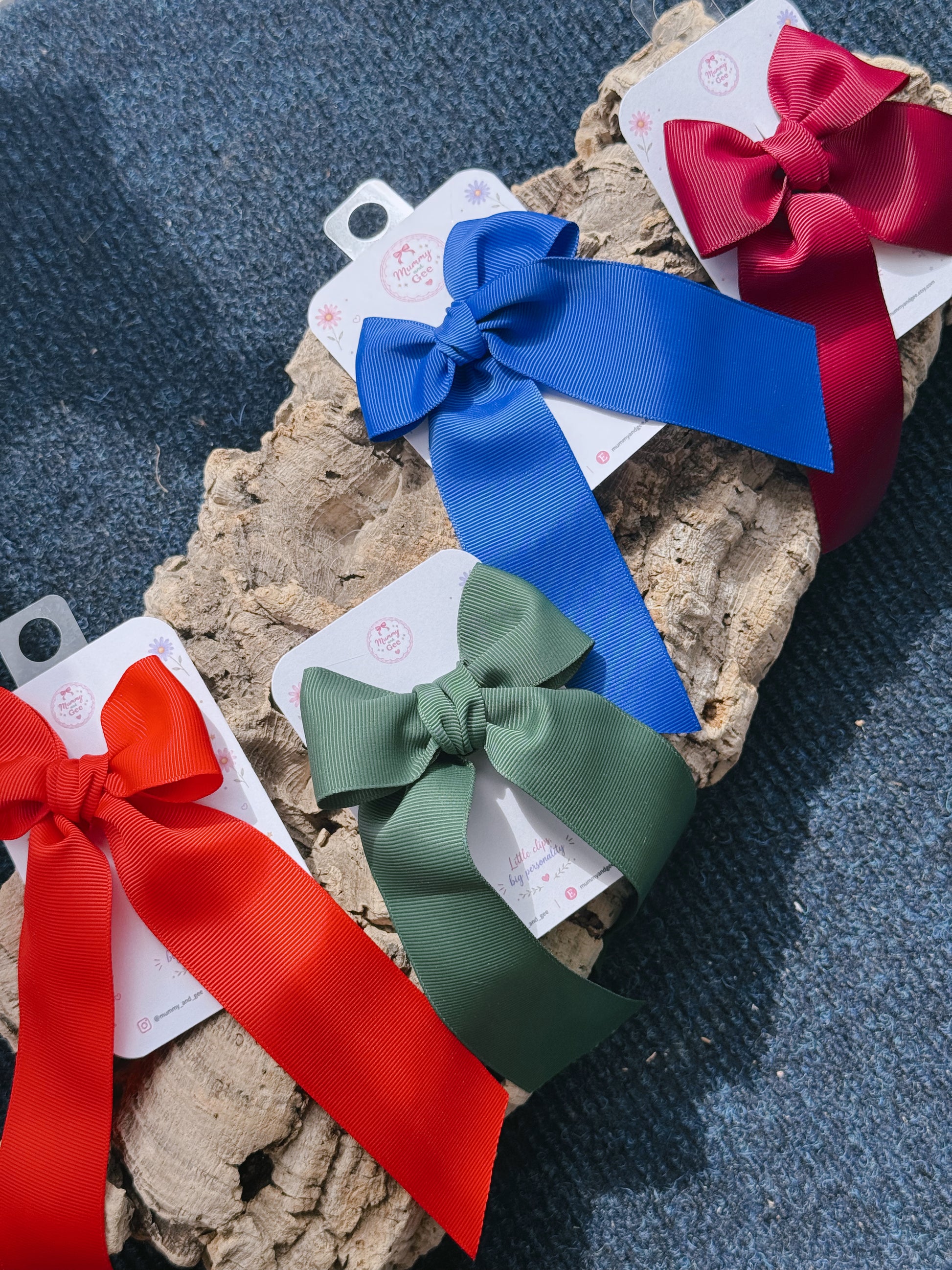 Three satin ribbon hair bows in red, blue, and green, each tied to a white paper tag and displayed on a piece of natural cork bark over a dark blue textured fabric background.